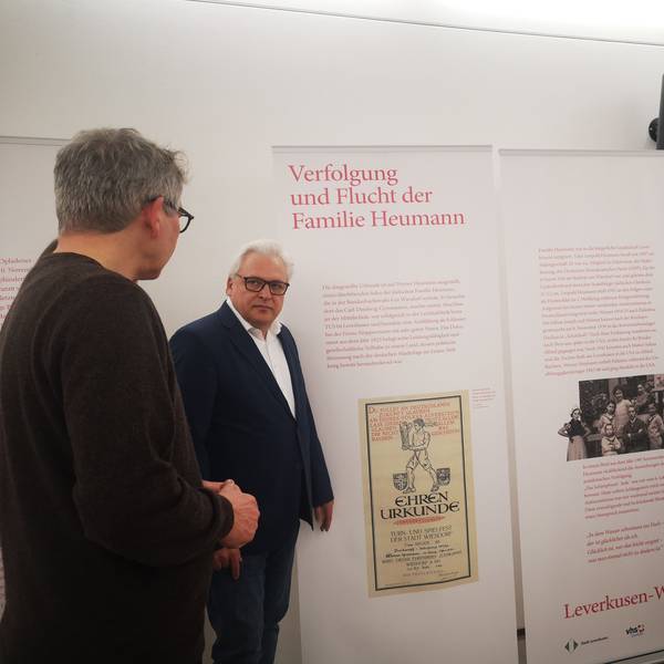 Mitarbeitende der VHS Leverkusen und der Stadt stehen vor einem Ausstellungsbanner mit dem Thema Verfolgung und Flucht der Familie Heumann