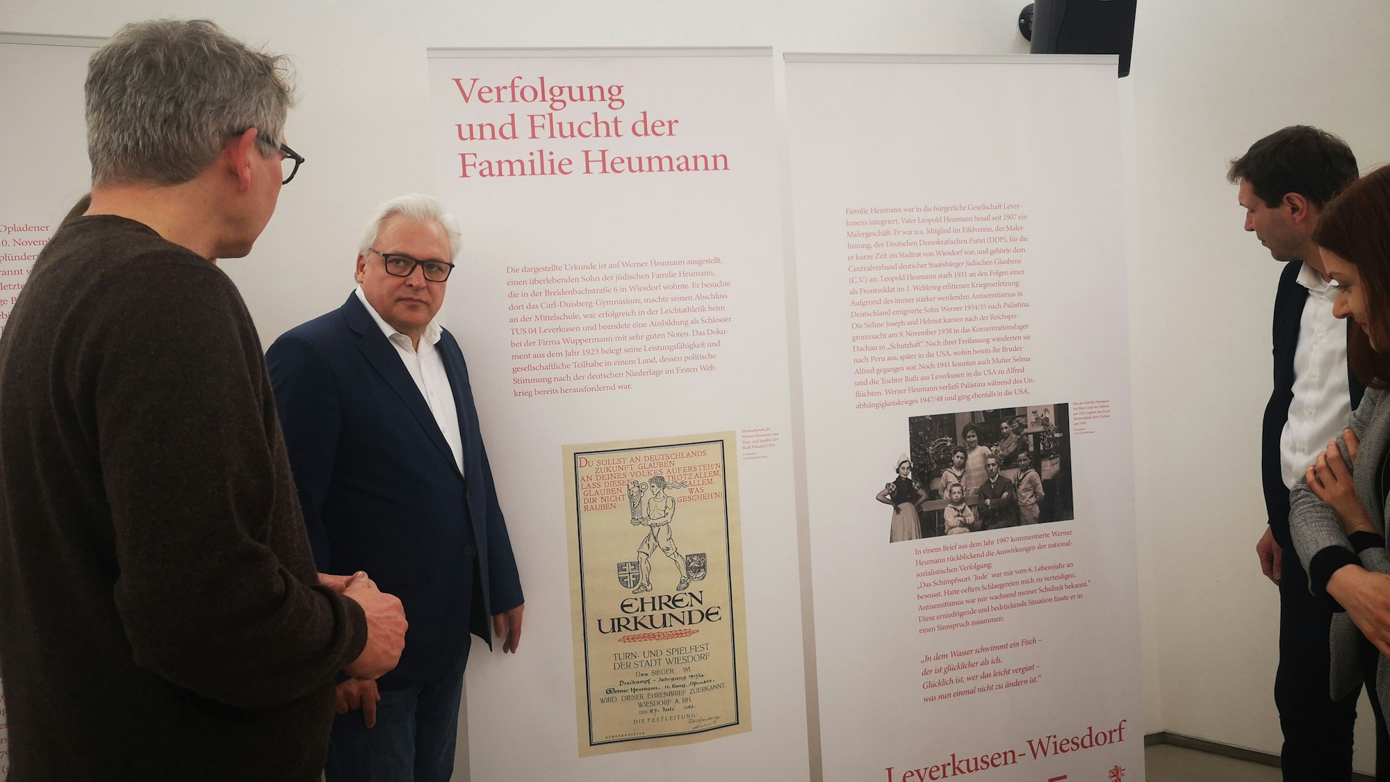 Mitarbeitende der VHS Leverkusen und der Stadt stehen vor einem Ausstellungsbanner mit dem Thema Verfolgung und Flucht der Familie Heumann