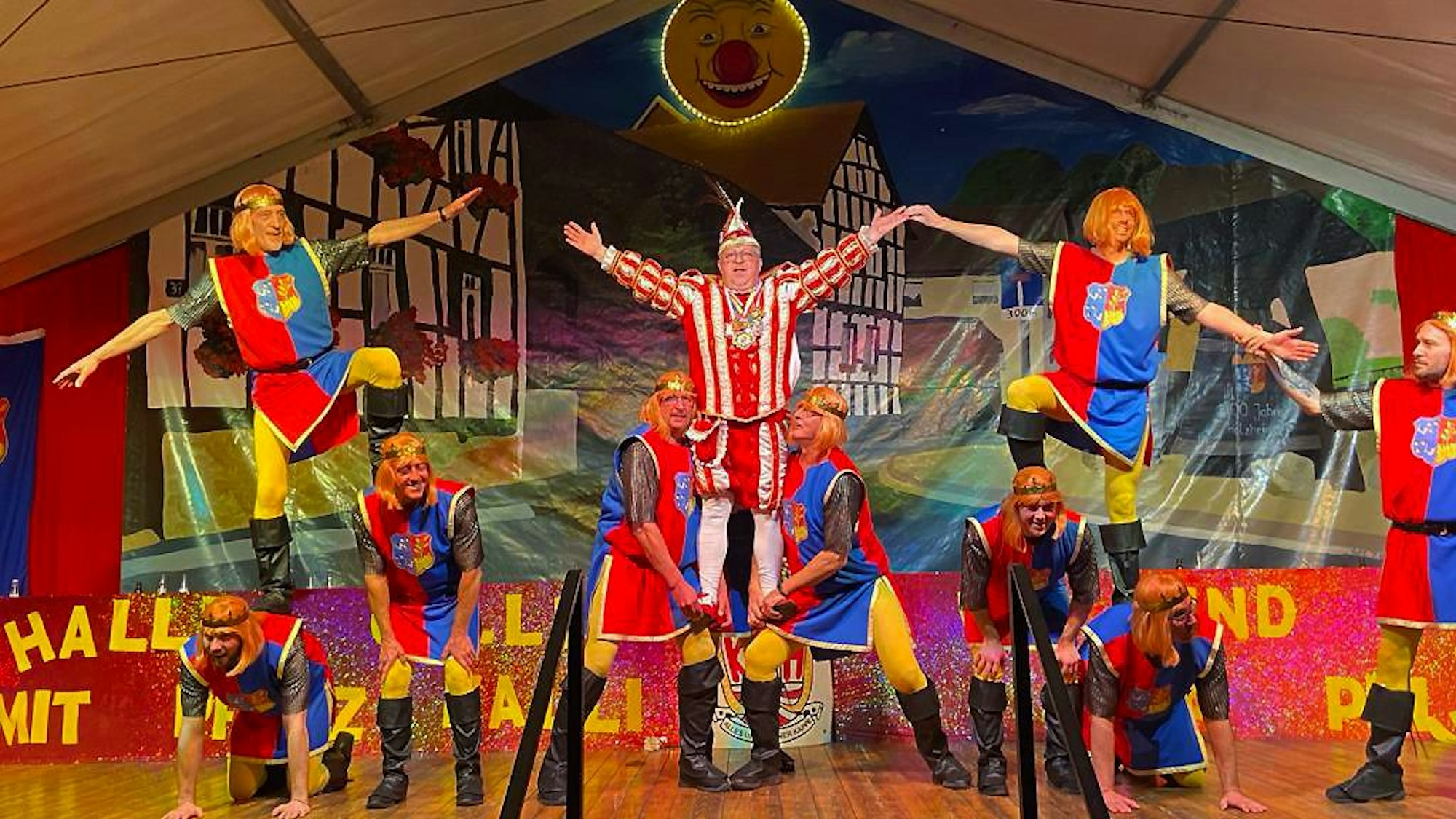 Tänzer des Männerballetts haben Prinz Kalli in eine artistische Nummer integriert. Zwei Mitglieder tragen ihn auf ihren Händen. Er streckt die Arme nach oben.