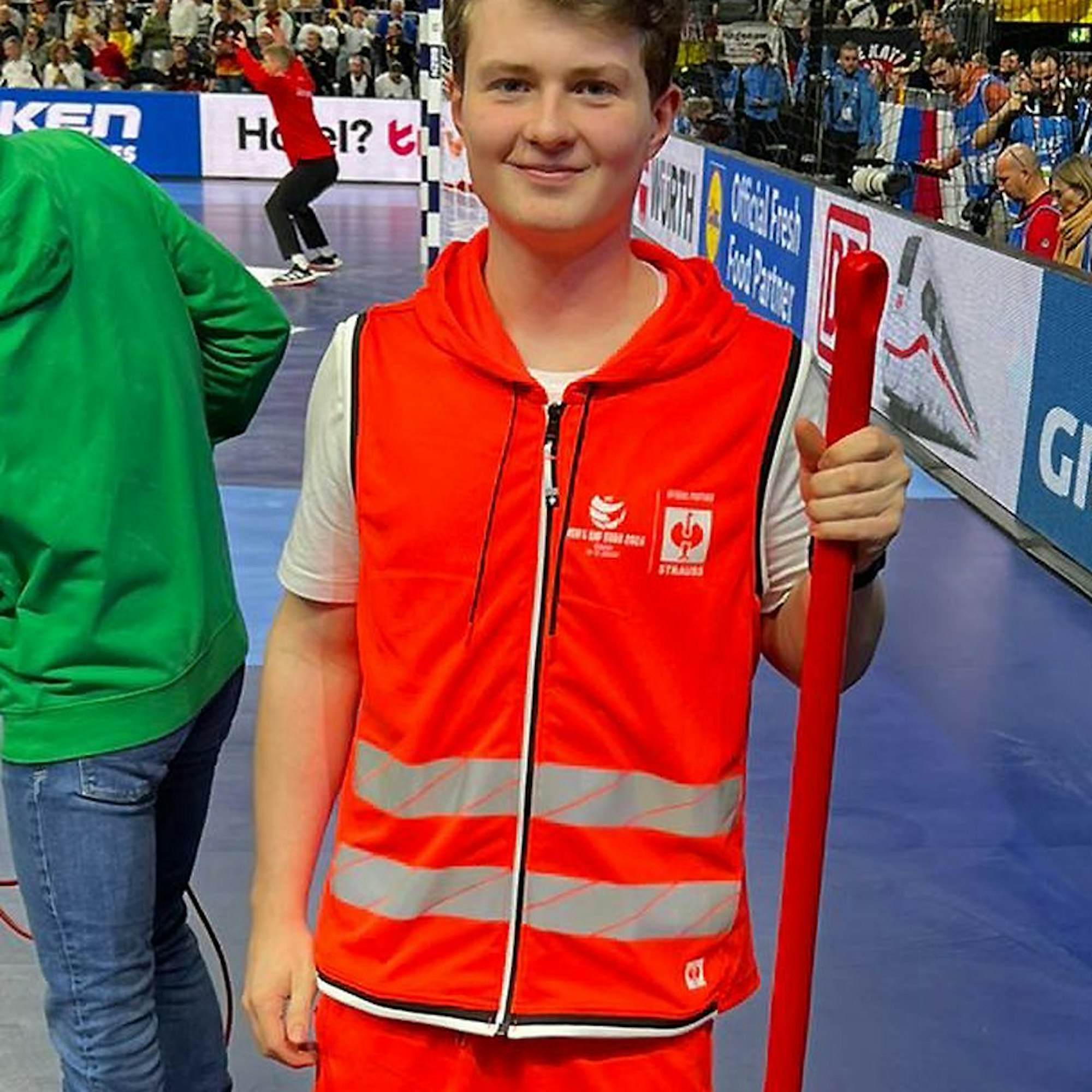 Ein junger Mann in Weste und kurzer Sporthose in leuchtend-orange, der einen Wischmop in der Hand hält.