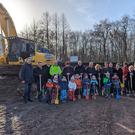 Eine Gruppe von Kindern mit Schaufeln und Erwachsenen mit Spaten auf einer Baustelle.