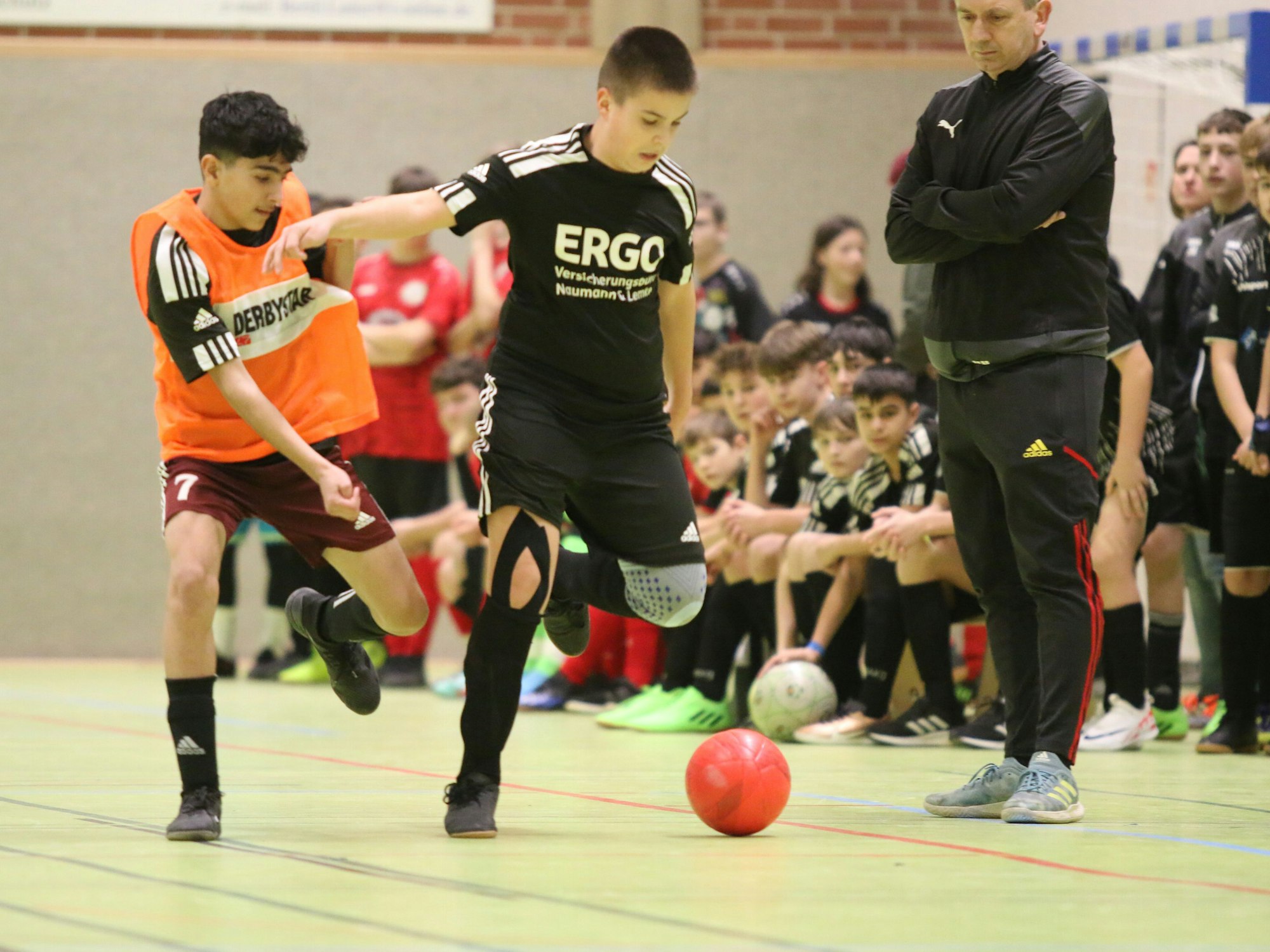 Zweikampf zwischen zwei Jugendfußballern. Einer von ihnen trägt ein orangefarbenes Leibchen. Der Trainer eines Teams steht direkt neben den Spielern.