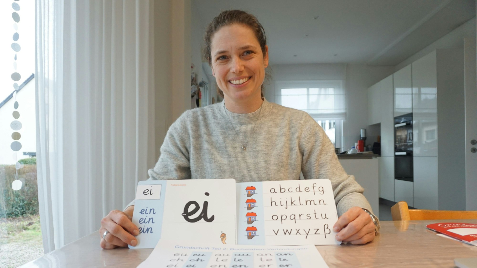 Eine Frau sitzt an einem Tisch und hält zwei Papierblätter mit handgeschriebenen Buchstaben in den Händen.