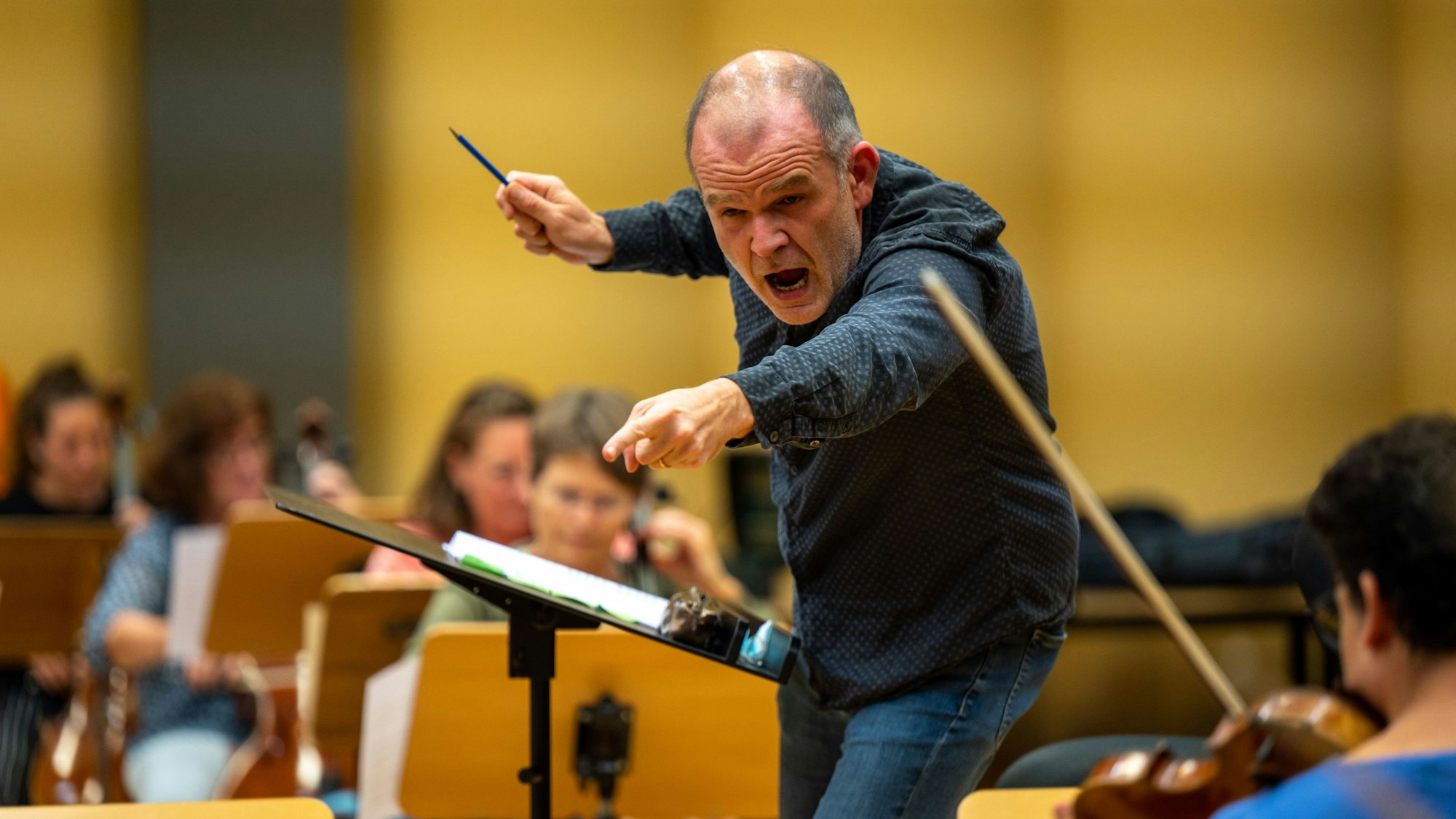 Der Dirigent macht energische Gesten in Richtung des Orchesters. Er hält einen Stift in der Hand.