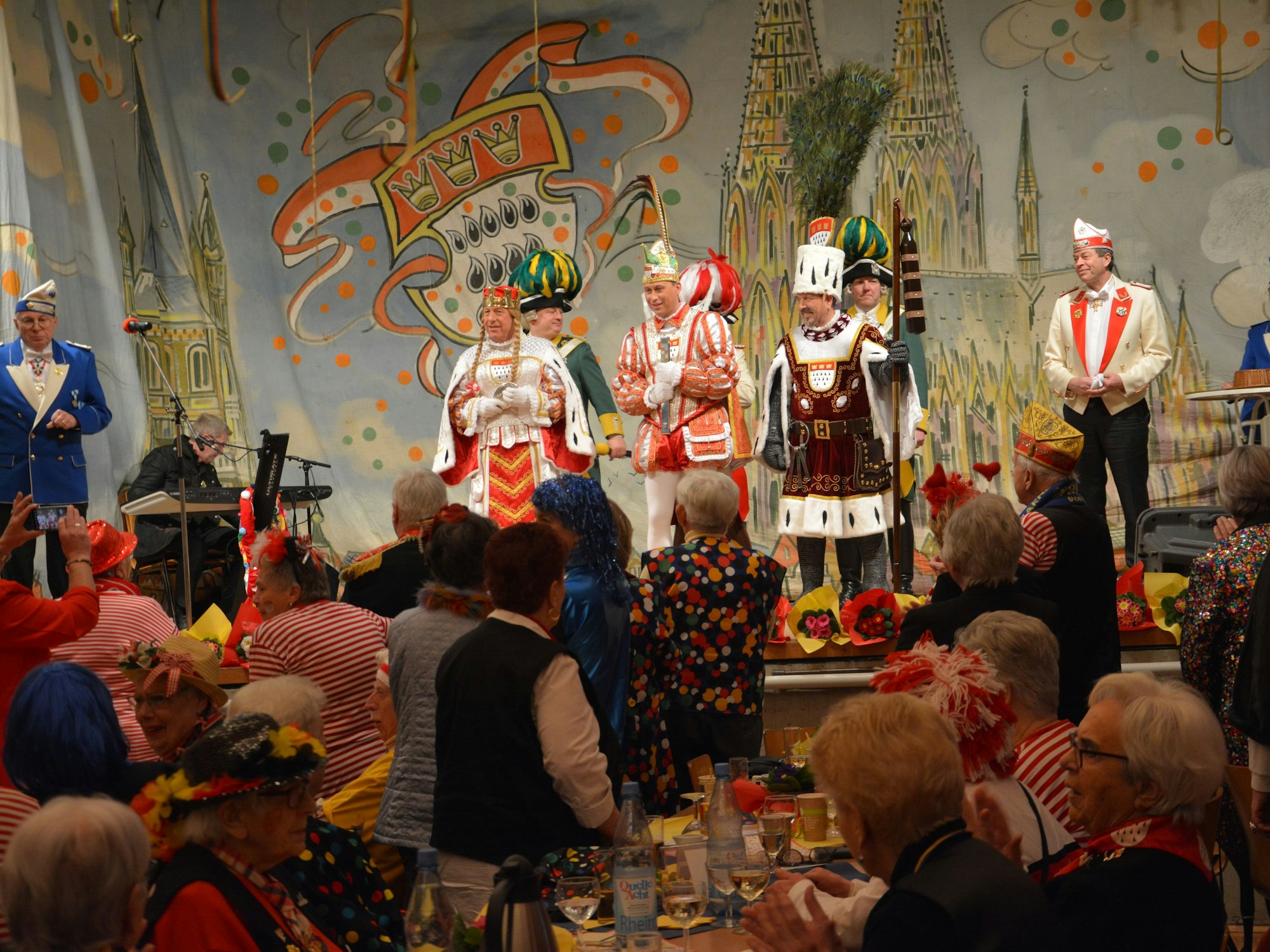 Blick in den Pfarrsaal, das Dreigestirn auf der Bühne, davor ältere Menschen.