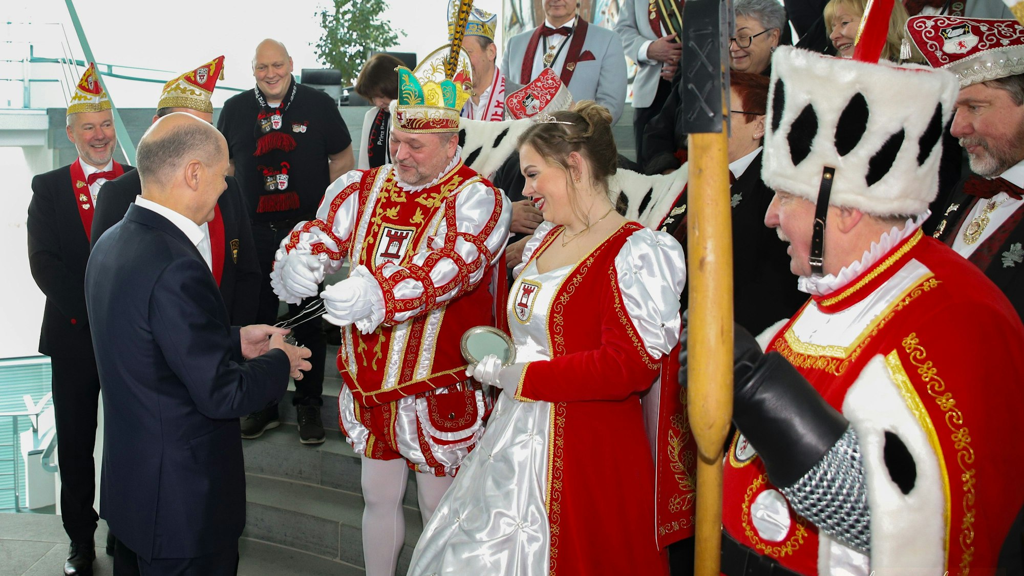 Mann im dunkelblauen Anzug, der einen Gegenstand von einem Mann in rot-weißer Karnevalstracht entgegen nimmt