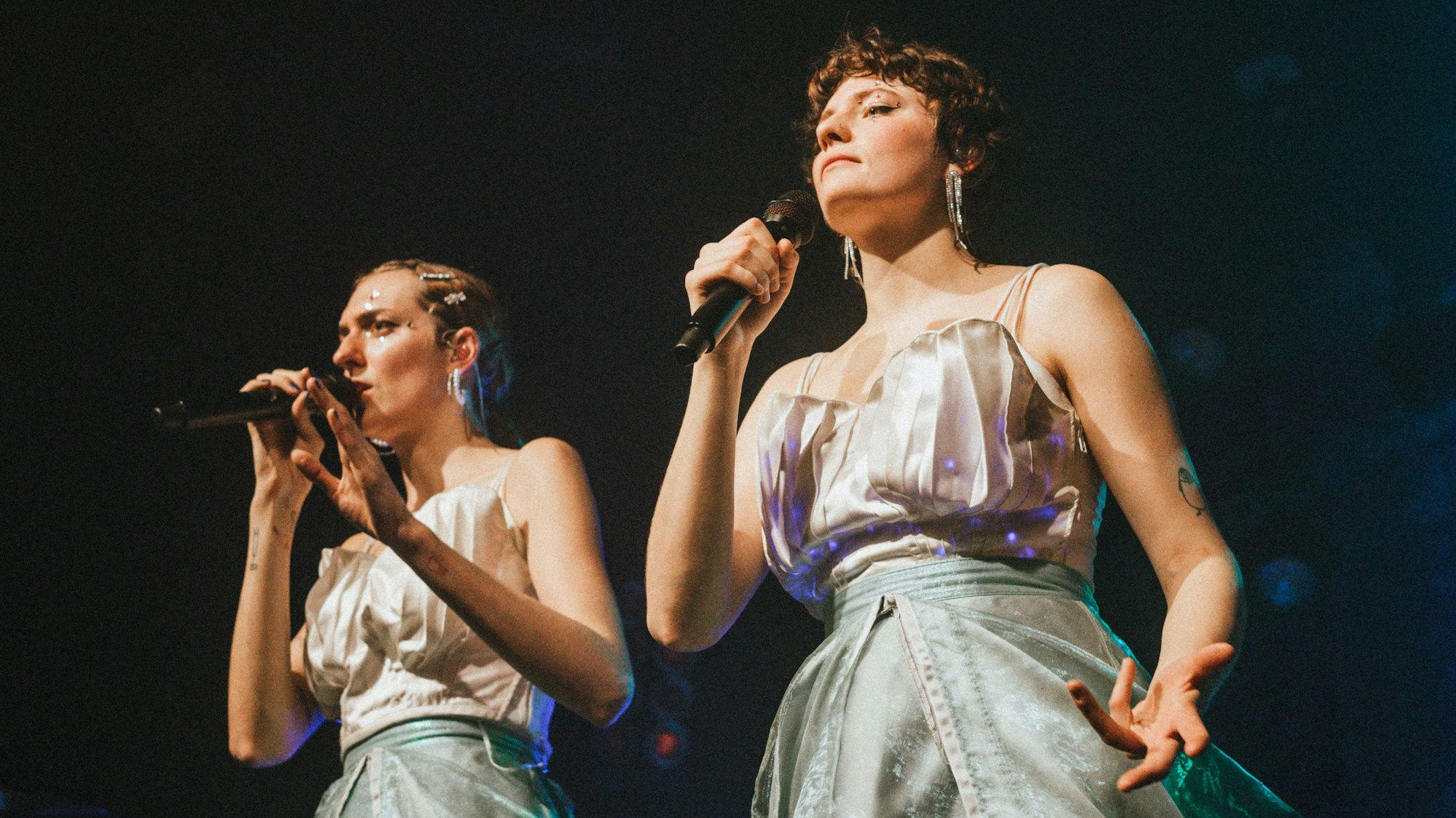 Zwei Sängerinnen in weißen Kleidern mit Glitzer im Gesicht.