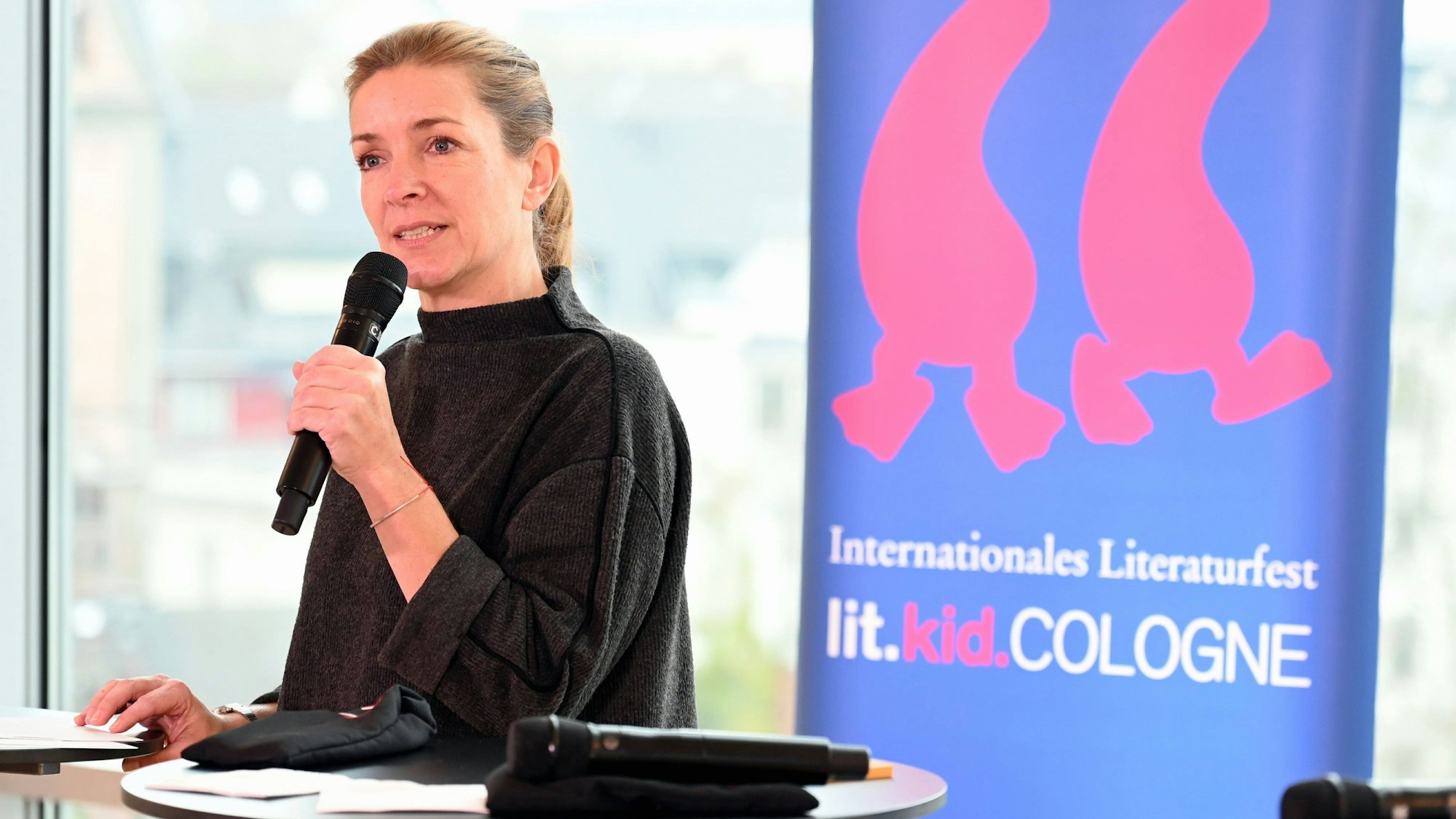 Angela Furtkamp spricht in ein Mikrofon, vor sich einige Stehtische. Hinter ihr steht ein Banner mit der Aufschrift: Internationales Literaturfest. lit.kid.COLOGNE.