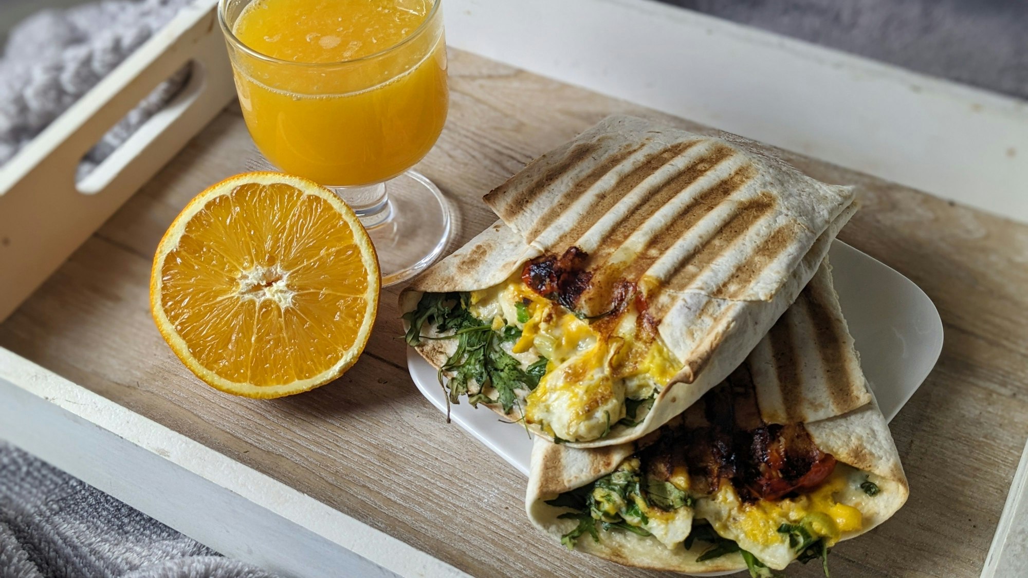 Zwei Weizentortillas liegen auf einem Teller, der neben einem Glas Orangensaft und einer halben Orange auf einem Tablett steht.
