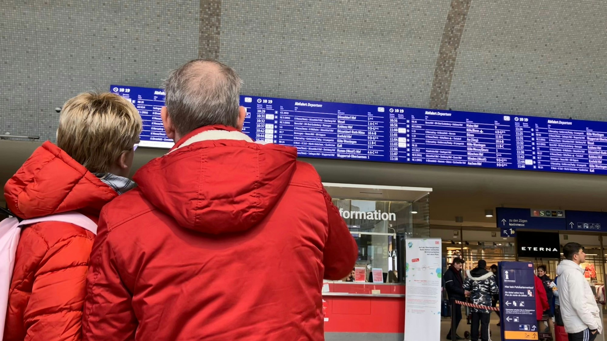 Ein Paar in Winterjacken sucht auf der Anzeigetafel im Kölner Hauptbahnhof nach einem Zug.