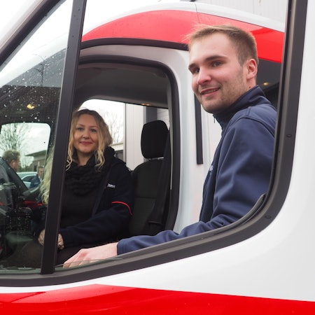 Eine Frau sitzt am Steuer eines Autos, ein Mann steht an der Tür.