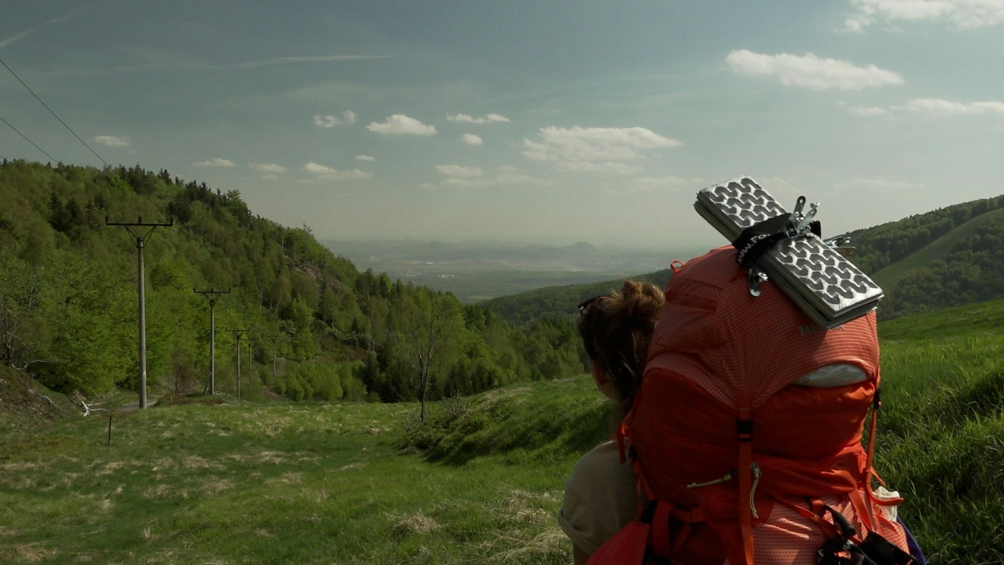 Die Protagonistin hat einen roten Rucksack und steht vorne im Bild. Sie schaut auf eine Wald- und Wiesenlandschaft.