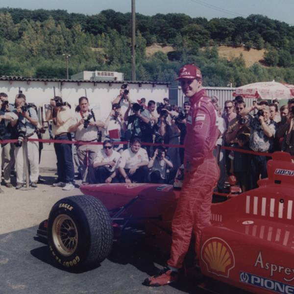 Auf dem Foto ist der Rennfahrer Michael Schumacher vor einem roten Rennwagen zu sehen.