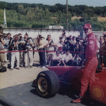 Auf dem Foto ist der Rennfahrer Michael Schumacher vor einem roten Rennwagen zu sehen.