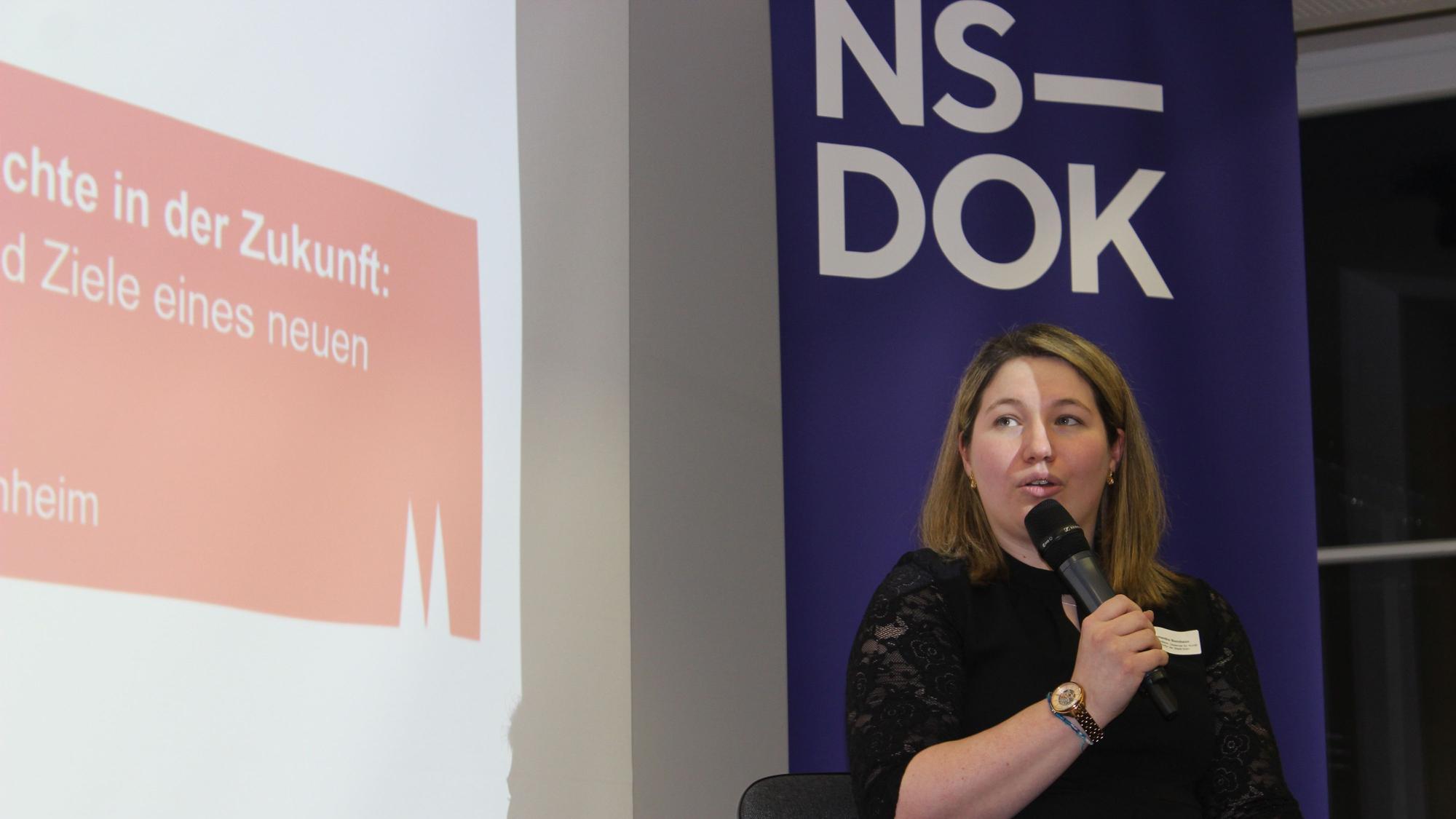 Frau im schwarzen Kleid, die vor einem Plakat mit der Aufschrift „NS-Dok“ in ein Mikro spricht.