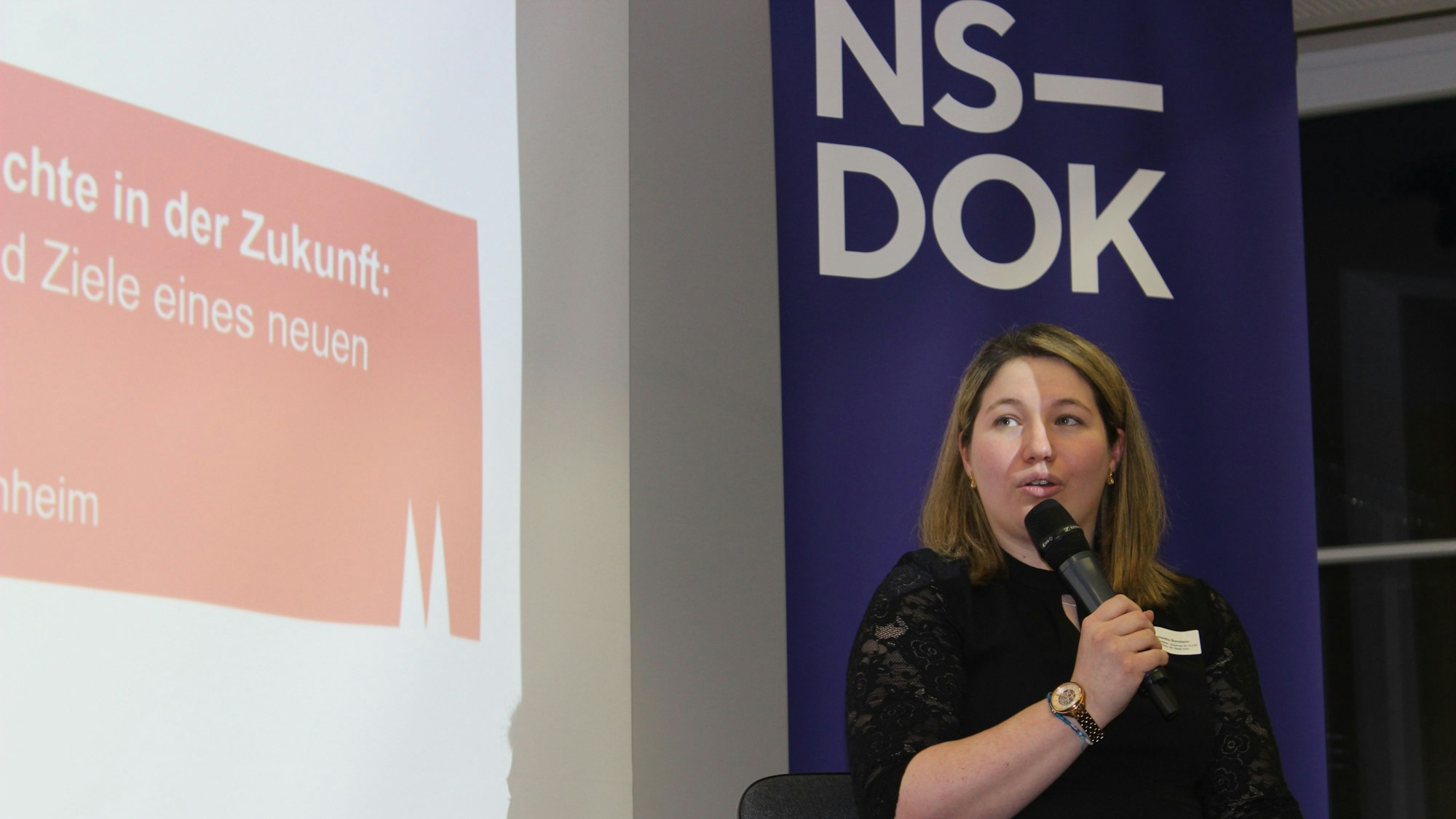 Frau im schwarzen Kleid, die vor einem Plakat mit der Aufschrift „NS-Dok“ in ein Mikro spricht.