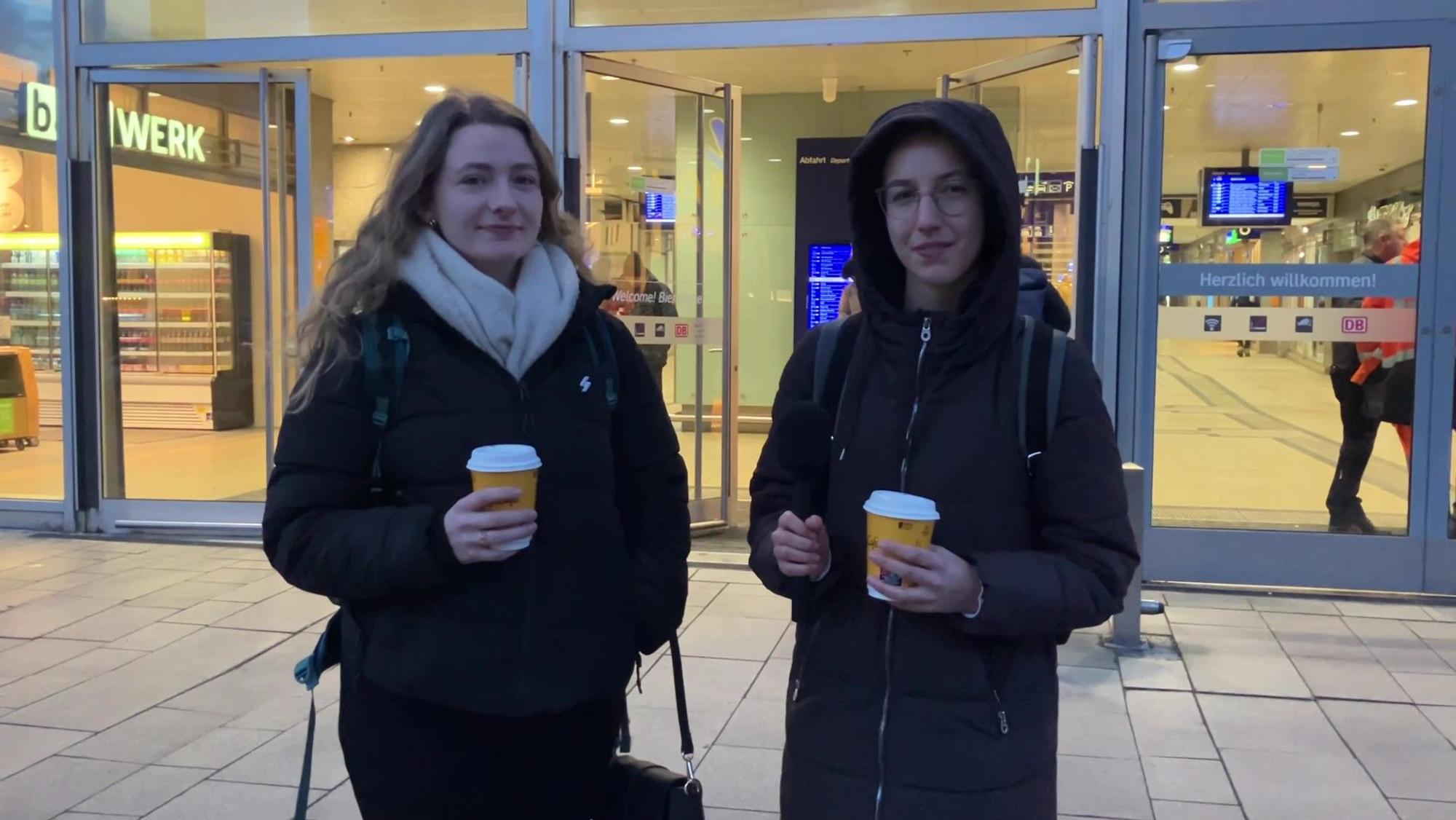 Zwei junge Frauen stehen mit Kaffeebechern in den Händen vor dem Kölner Hauptbahnhof.