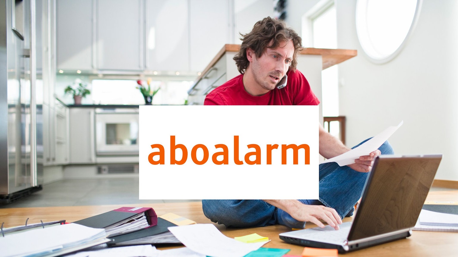Mann sitzt an seinem Laptop auf dem Boden und um ihn herum liegen Papierstapel. Logo von Aboalarm in der Bildmitte