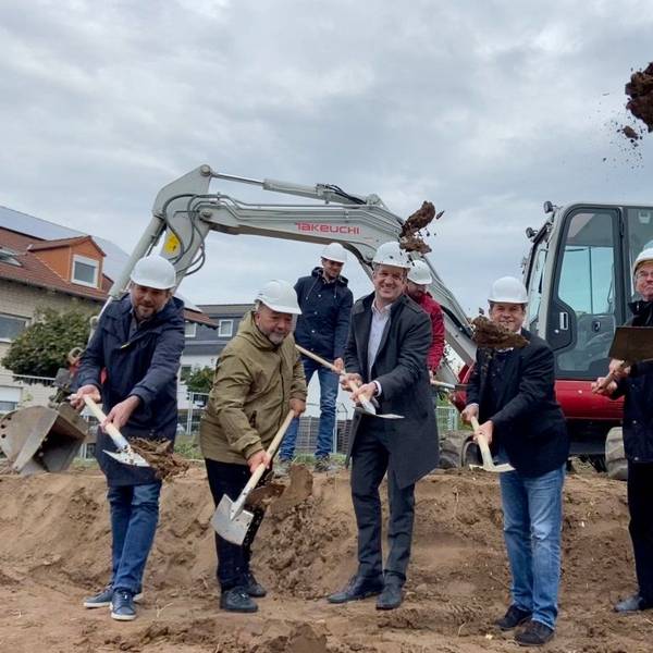 Fünf Männer stehen auf einem Sandhaufen, sie schaufeln Sand, im Hintergrund steht ein Bagger.