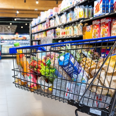 Ein voller Einkaufswagen biegt in einen Gang im Supermarkt zwischen zwei Regalen.