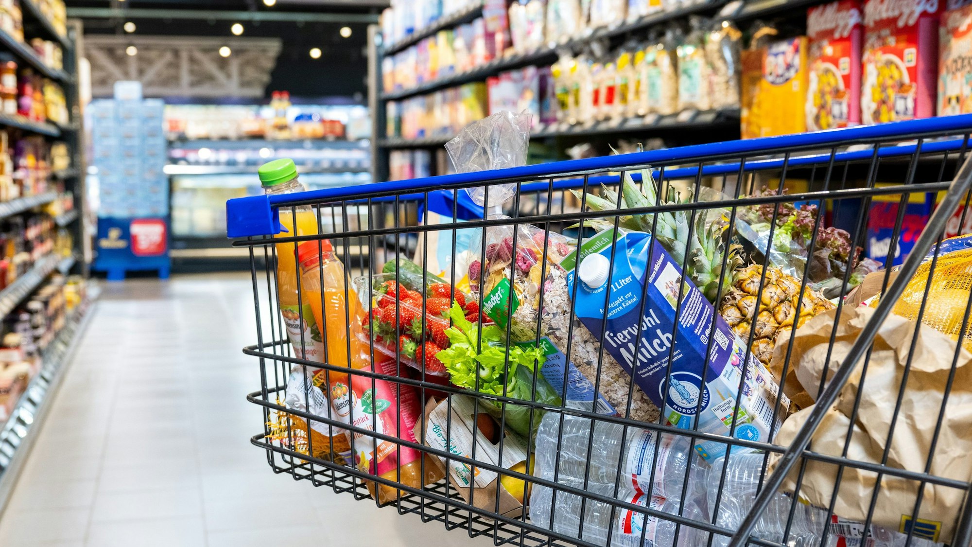 Ein voller Einkaufswagen biegt in einen Gang im Supermarkt zwischen zwei Regalen.