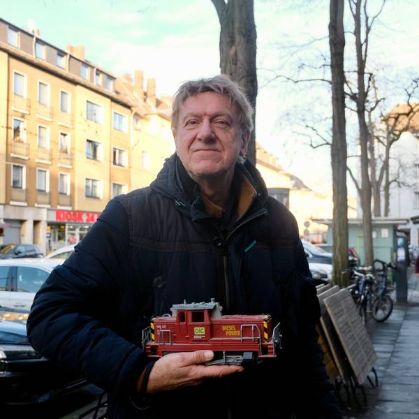 Ein Mann steht auf einem Bürgersteig an einer belebten Straße. Er hält eine Modell-Lok in der Hand.