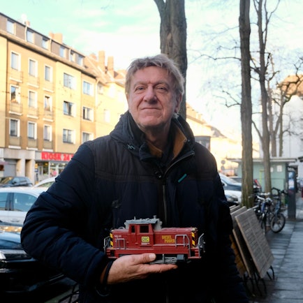 Ein Mann steht auf einem Bürgersteig an einer belebten Straße. Er hält eine Modell-Lok in der Hand.