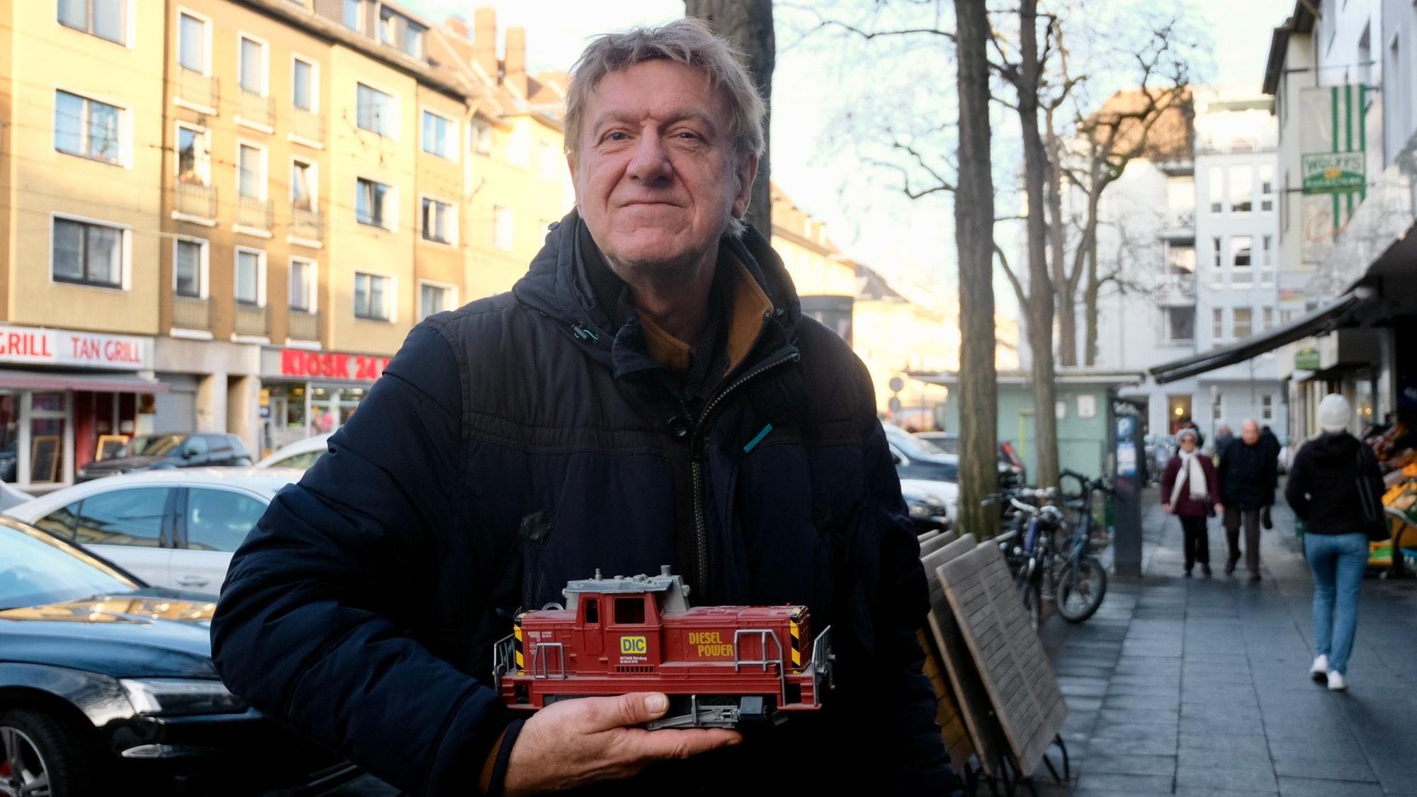 Ein Mann steht auf einem Bürgersteig an einer belebten Straße. Er hält eine Modell-Lok in der Hand.