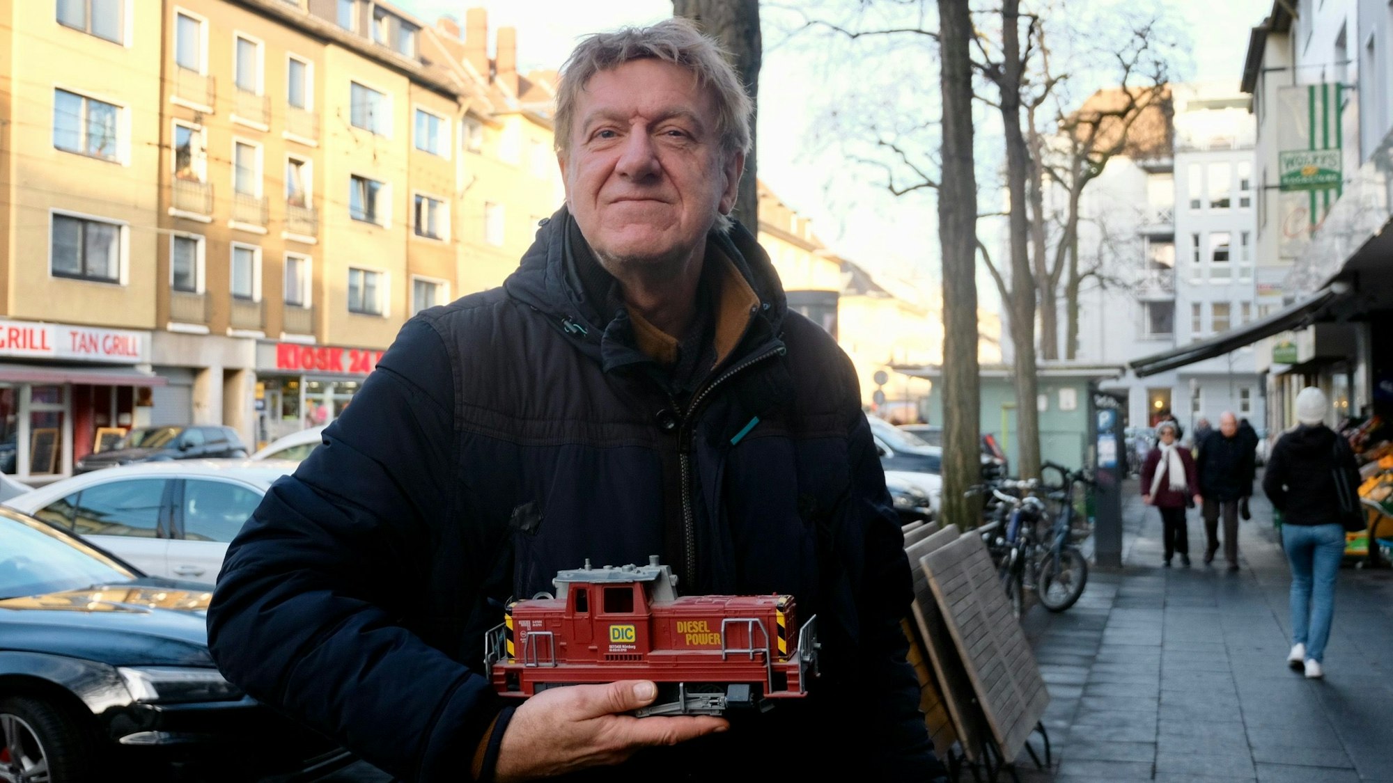 Ein Mann steht auf einem Bürgersteig an einer belebten Straße. Er hält eine Modell-Lok in der Hand.