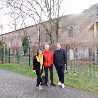 Eine Frau und zwei Männer stehen vor einem massiven zweigeschossigen Klinkerbau.