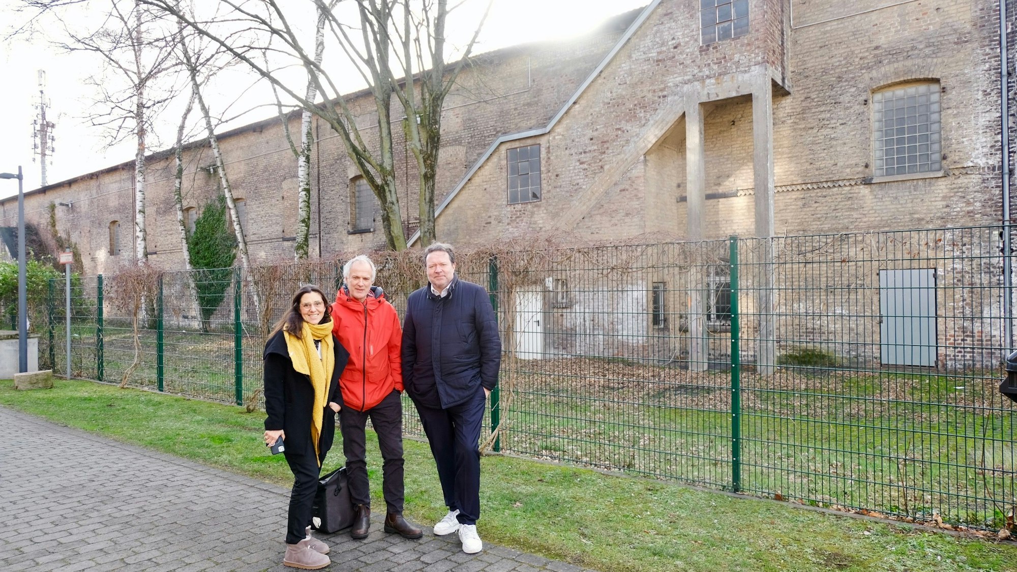 Eine Frau und zwei Männer stehen vor einem massiven zweigeschossigen Klinkerbau.