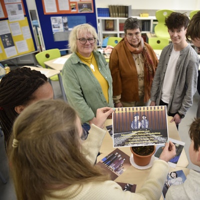 Schülerinnen und Schüler stehen im Kreis um einen Tisch mit verschiedenen Zeichnungen. Ein Mädchen hält ein Plakat in die Kamera.
