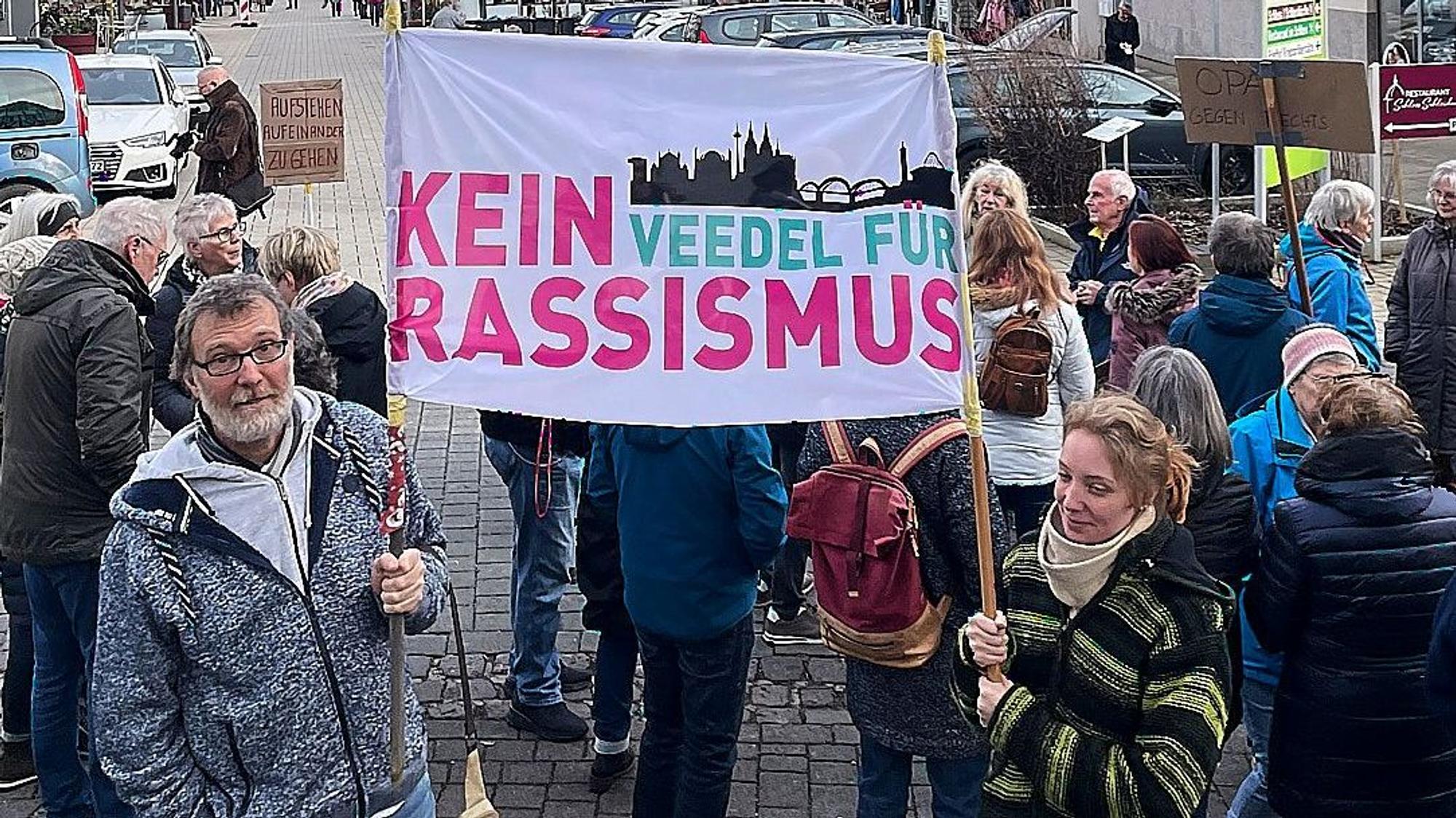 Vater und Tochter, Bernd und Maxi Sommer, halten ein weißes Banner mit der Aufschrift „Kein Veedel für Rassismus“ in pink und türkis sowie der Silhouette von Köln.