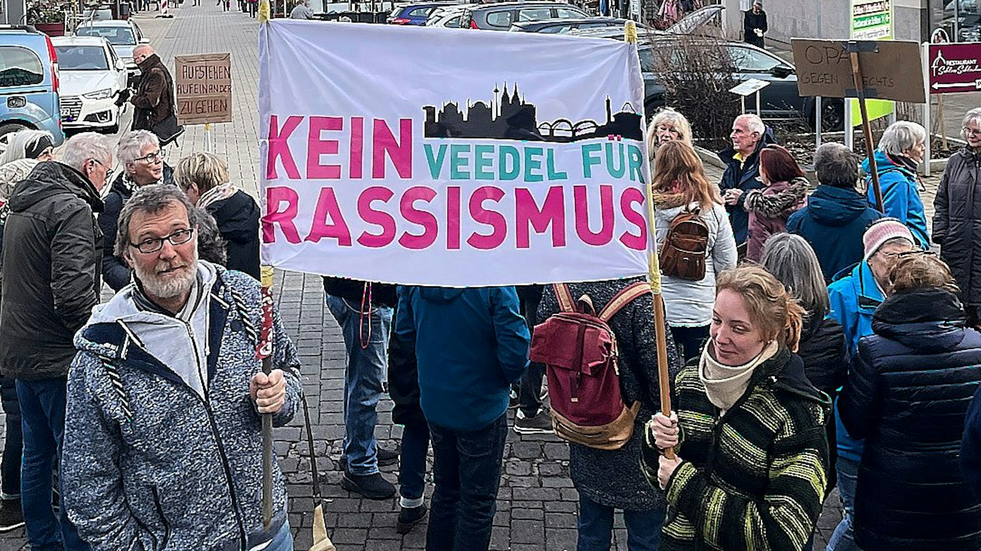 Vater und Tochter, Bernd und Maxi Sommer, halten ein weißes Banner mit der Aufschrift „Kein Veedel für Rassismus“ in pink und türkis sowie der Silhouette von Köln.