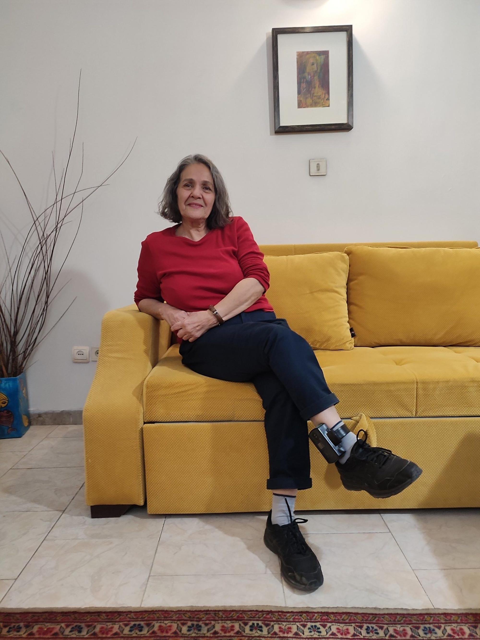 Nahid Taghavi sitzt auf einem gelben Sofa, am Fußgelenk ist eine elektronische Fußfessel zu sehen.