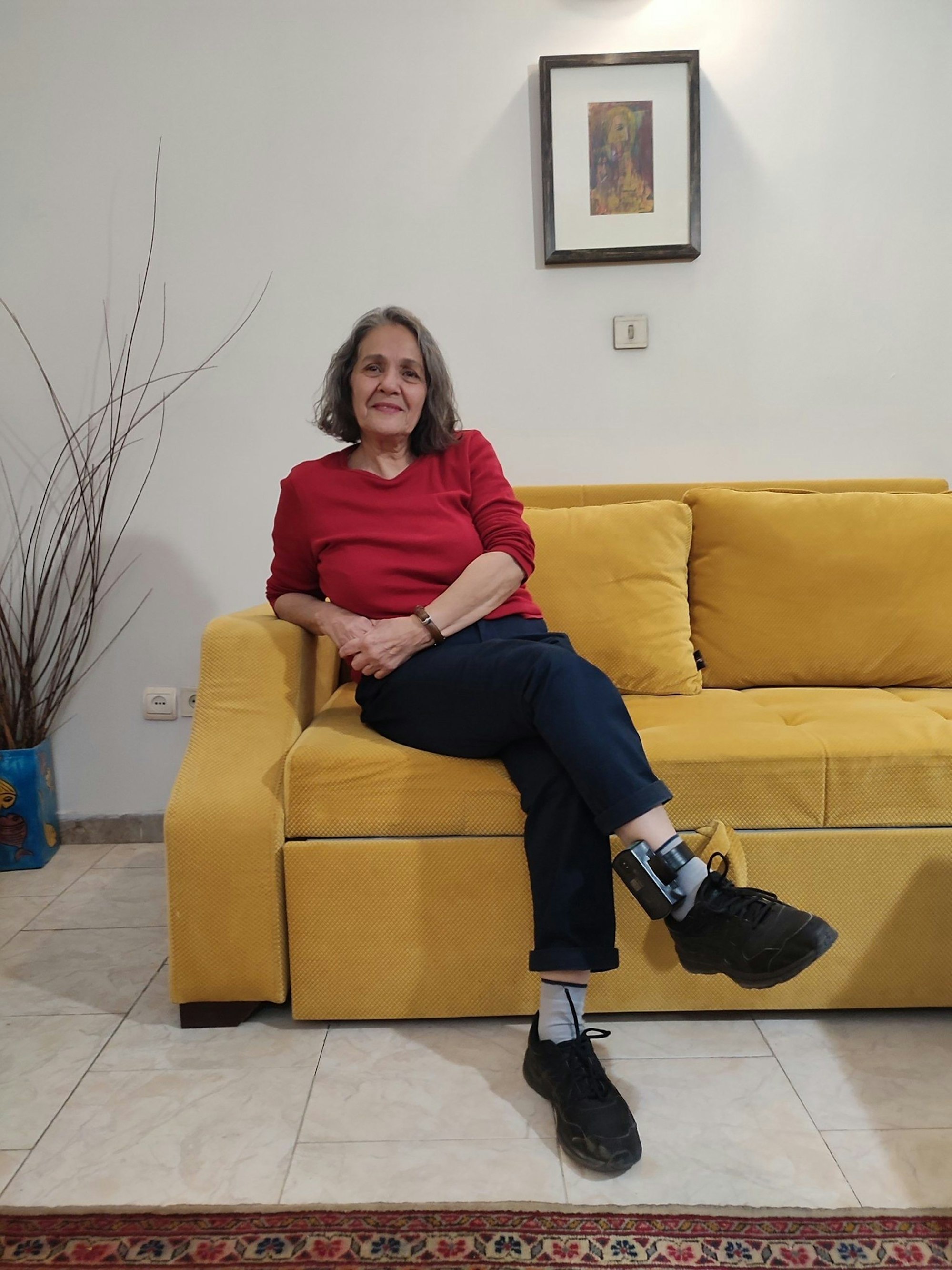 Nahid Taghavi sitzt auf einem gelben Sofa, am Fußgelenk ist eine elektronische Fußfessel zu sehen.