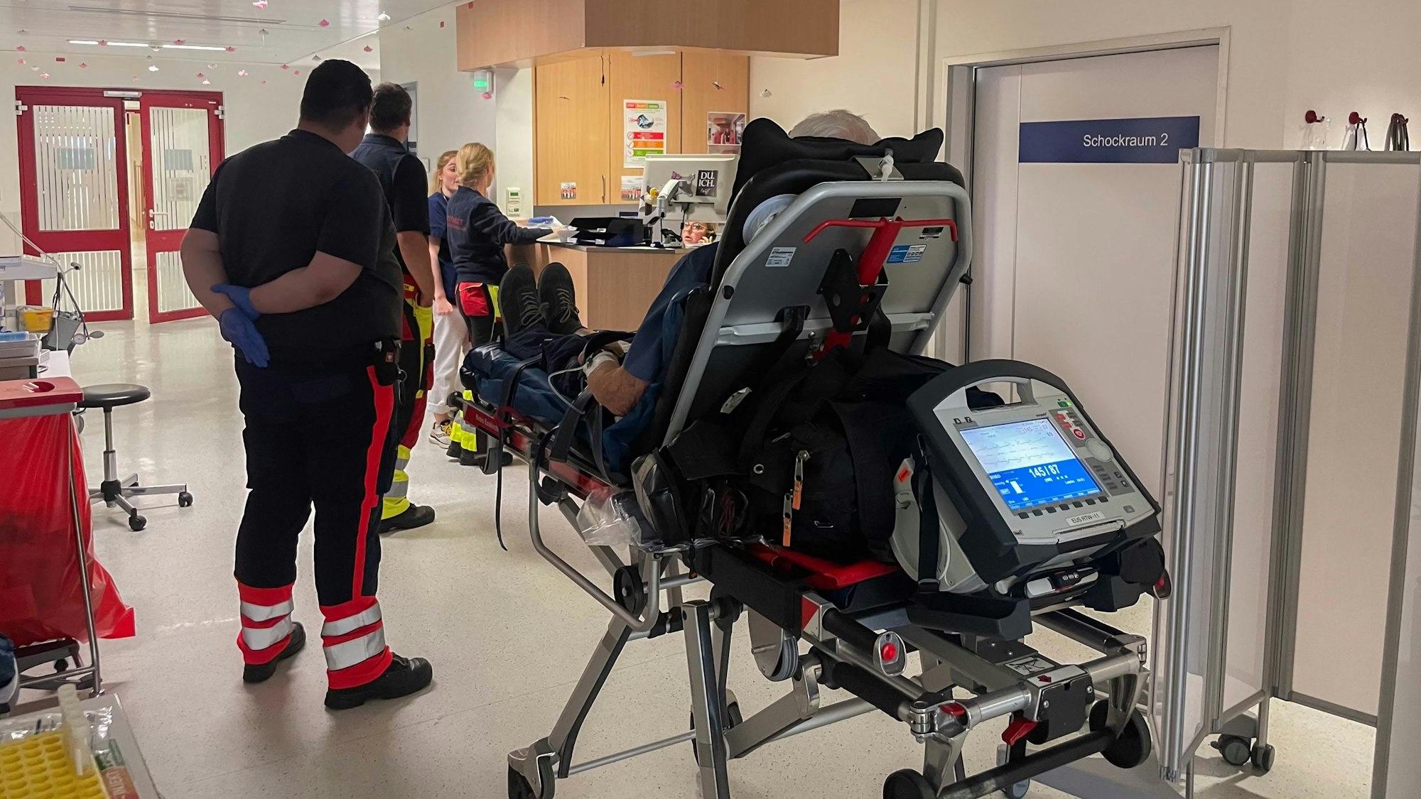 Das Bild zeigt eine Szene in der Notaufnahme. Ein Patient sitzt auf der Liege. Die Mitarbeiter des Rettungsdienstes warten an der Notaufnahme.