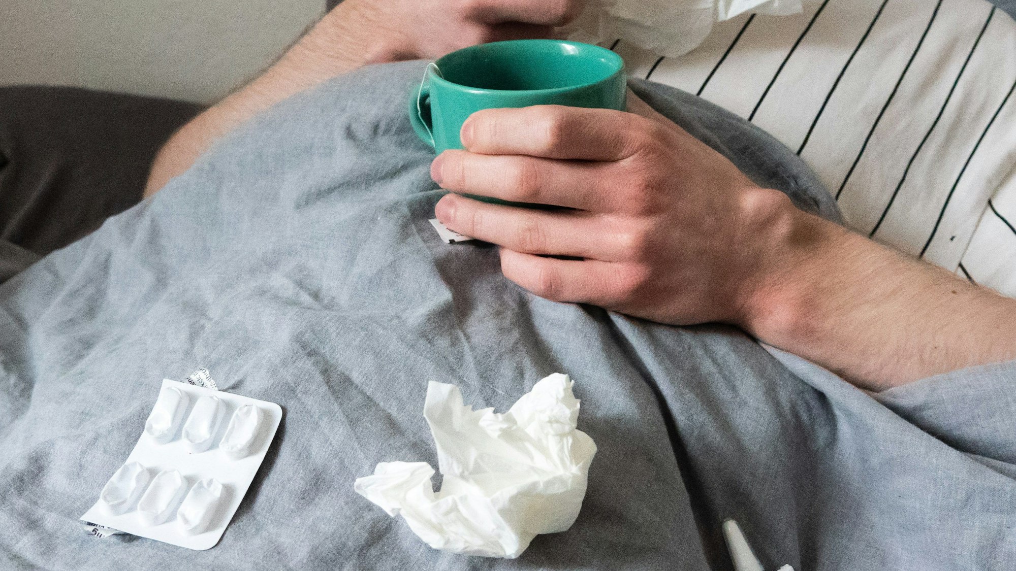 Ein Mann liegt mit Taschentüchern, Teetasse, Nasenspray und Tabletten im Bett (gestellte Szene).