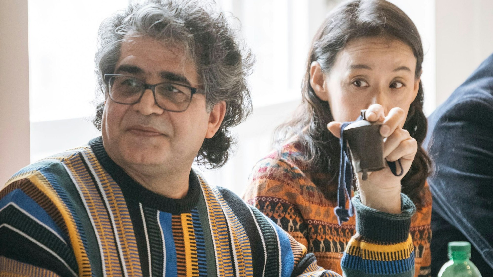 Ali Abdollahi trägt einen gestreiften, bunten Pullover. Er hält eine Schafsglocke aus seiner Kindheit hoch.