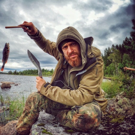 Zu sehen ist einer der Entwickler von „7 vs. Wild“, Fritz Meinecke, beim Angeln. Er sitzt an einem Fluss und hält einen Fisch in die Kamera.