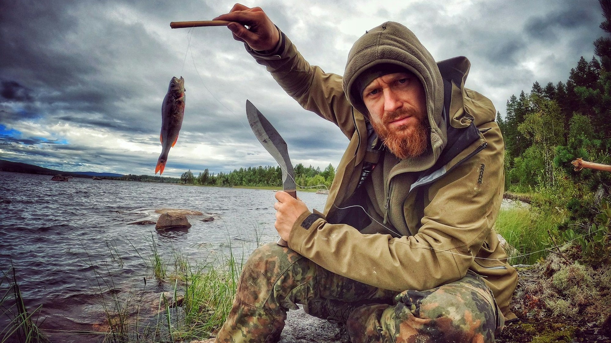 Zu sehen ist einer der Entwickler von „7 vs. Wild“, Fritz Meinecke, beim Angeln. Er sitzt an einem Fluss und hält einen Fisch in die Kamera.