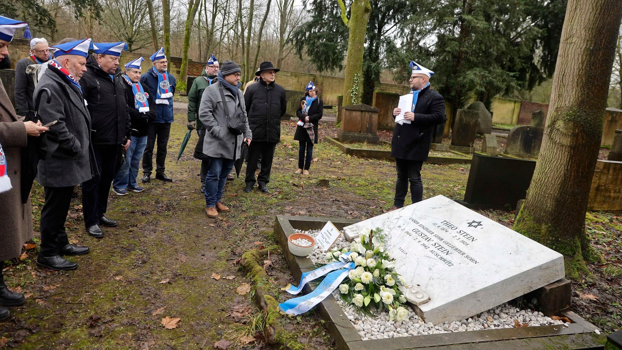 Eine Gruppe Menschen steht an einer Gedenktafel auf einem Friedhof.