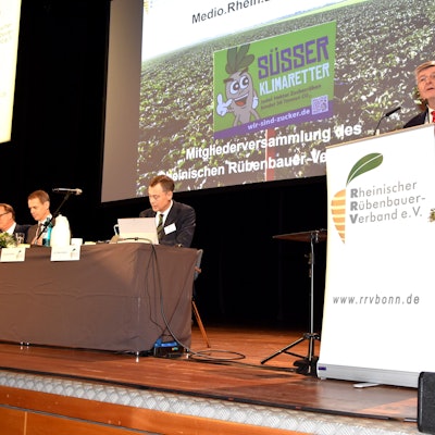 Ein Mann steht an einem Rednerpult auf einer Bühne, neben ihm sitzen Männer an einem Tisch.
