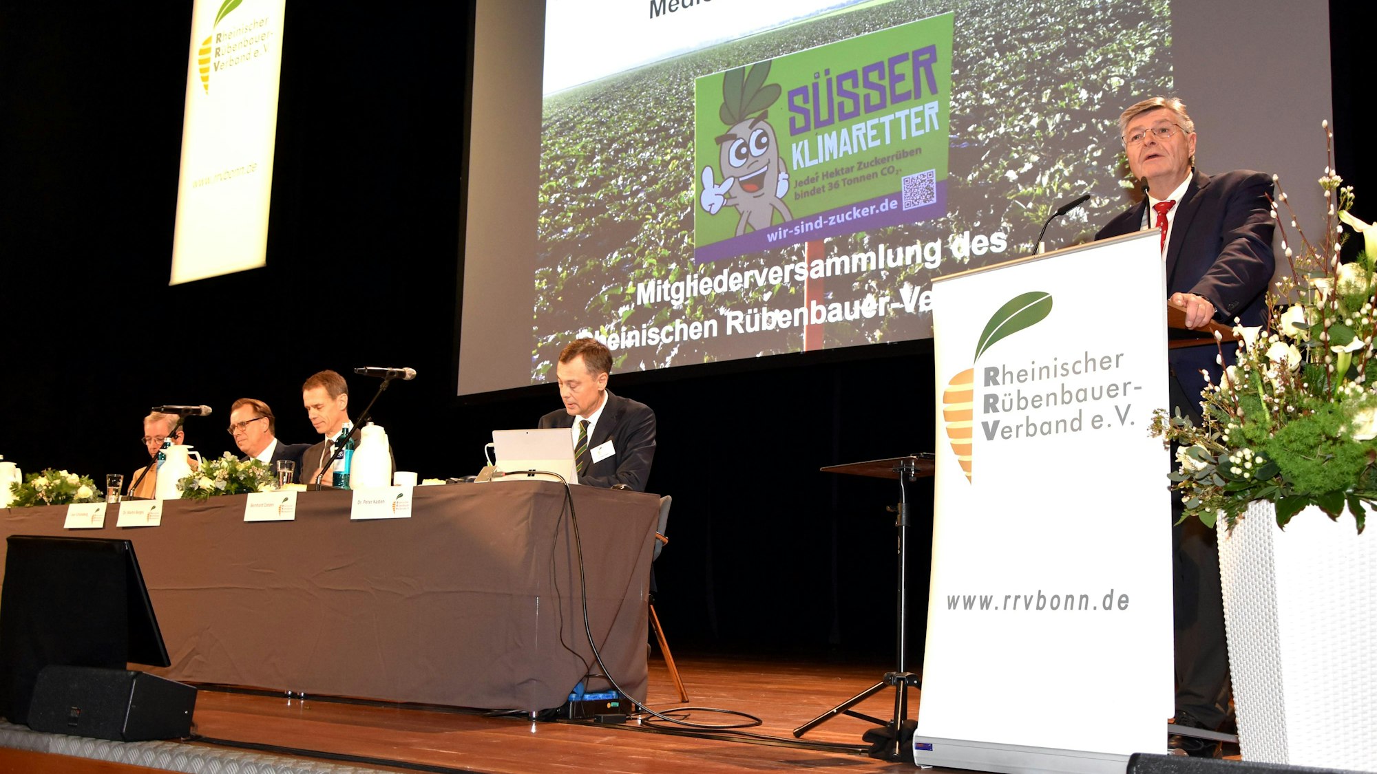 Ein Mann steht an einem Rednerpult auf einer Bühne, neben ihm sitzen Männer an einem Tisch.