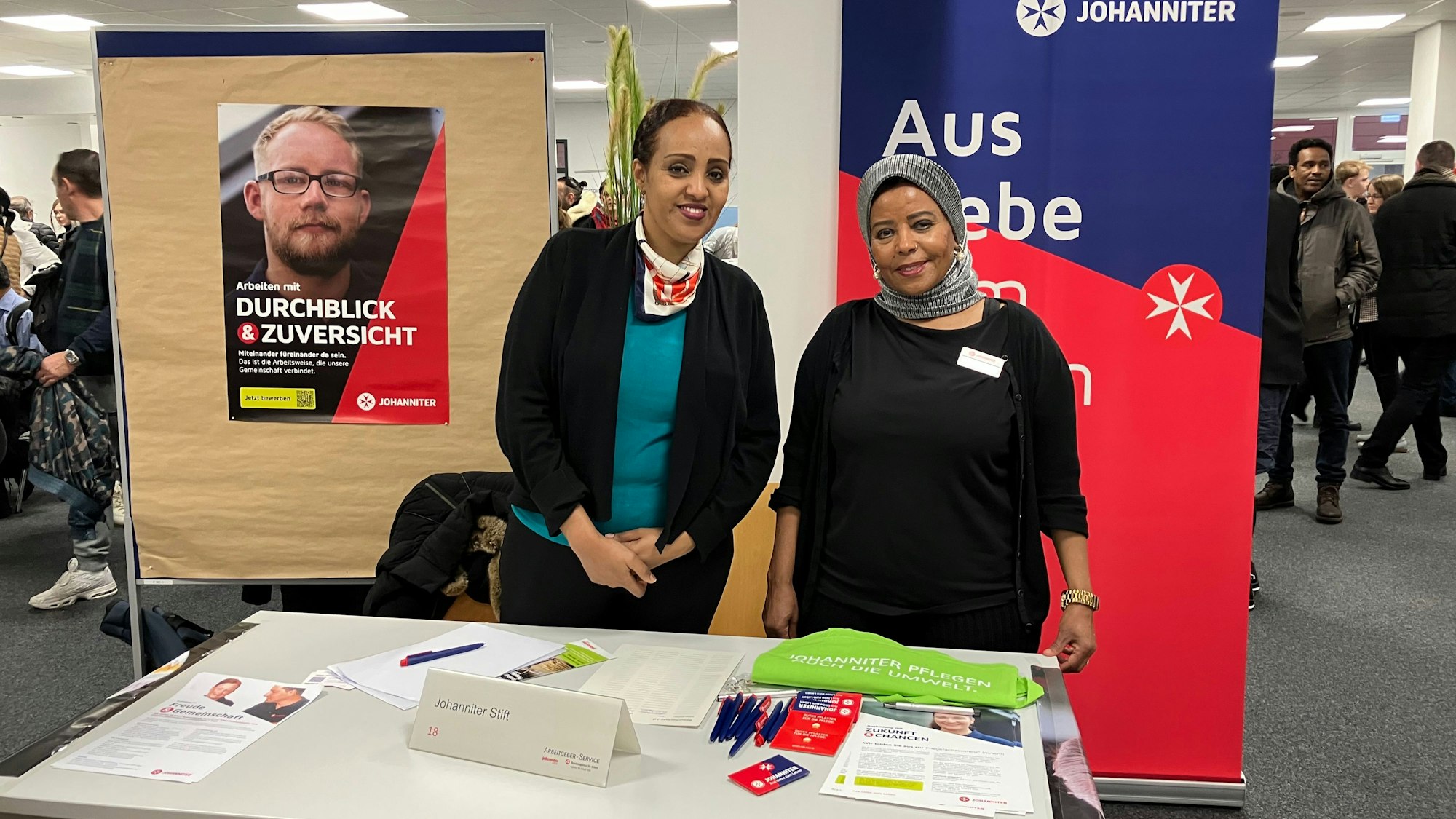 Najaha Abdurahman und ihre Kollegin stehen an einem Tisch mit Broschüren, hinter ihnen ein Plakat der Johanniter.