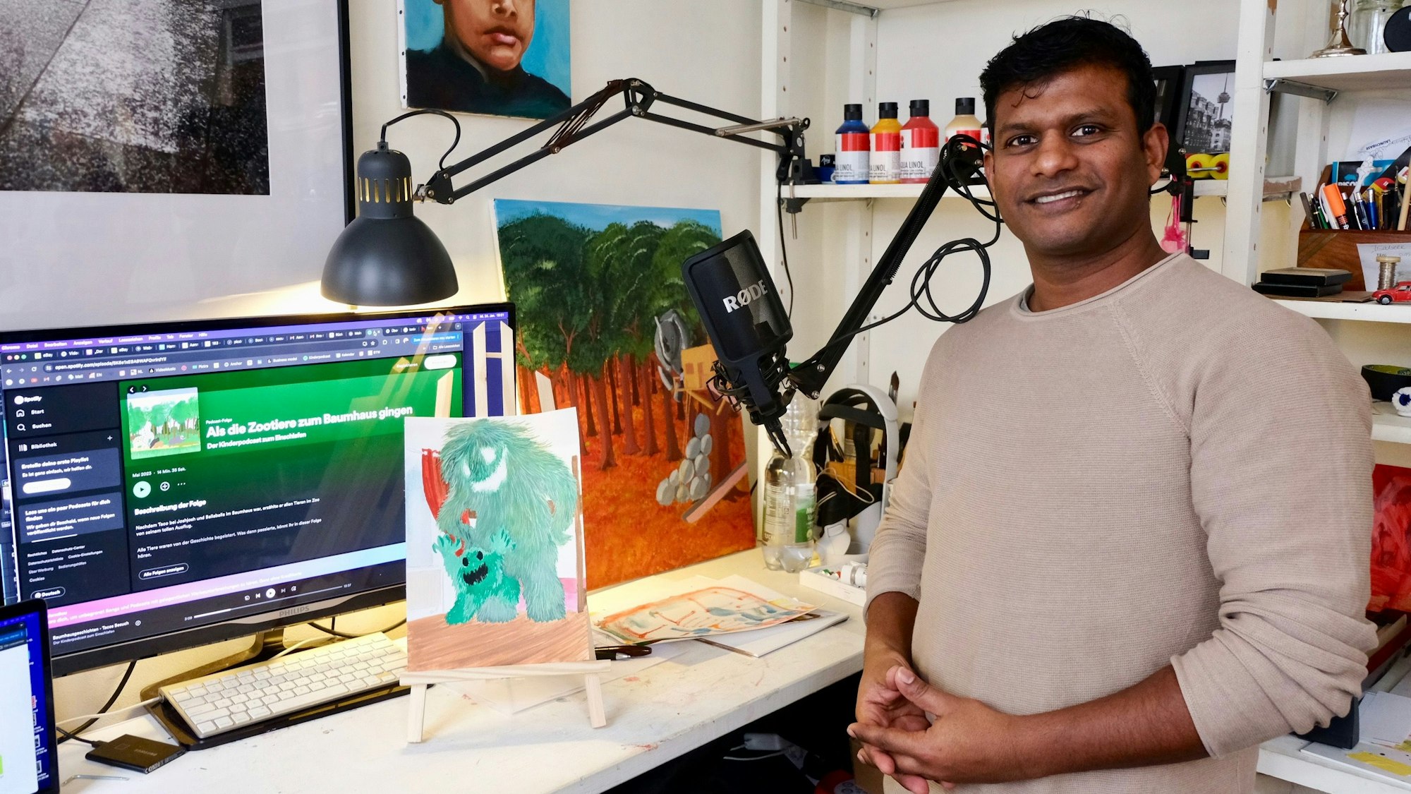 Ein Mann mit dunkler Hautfarbe und schwarzem Haar steht an einem Schreibtisch, auf dem ein großer Monitor steht, daneben ist eine Kinderzeichnung mit zwei Monstern auf einer Mini-Staffelei zu sehen.