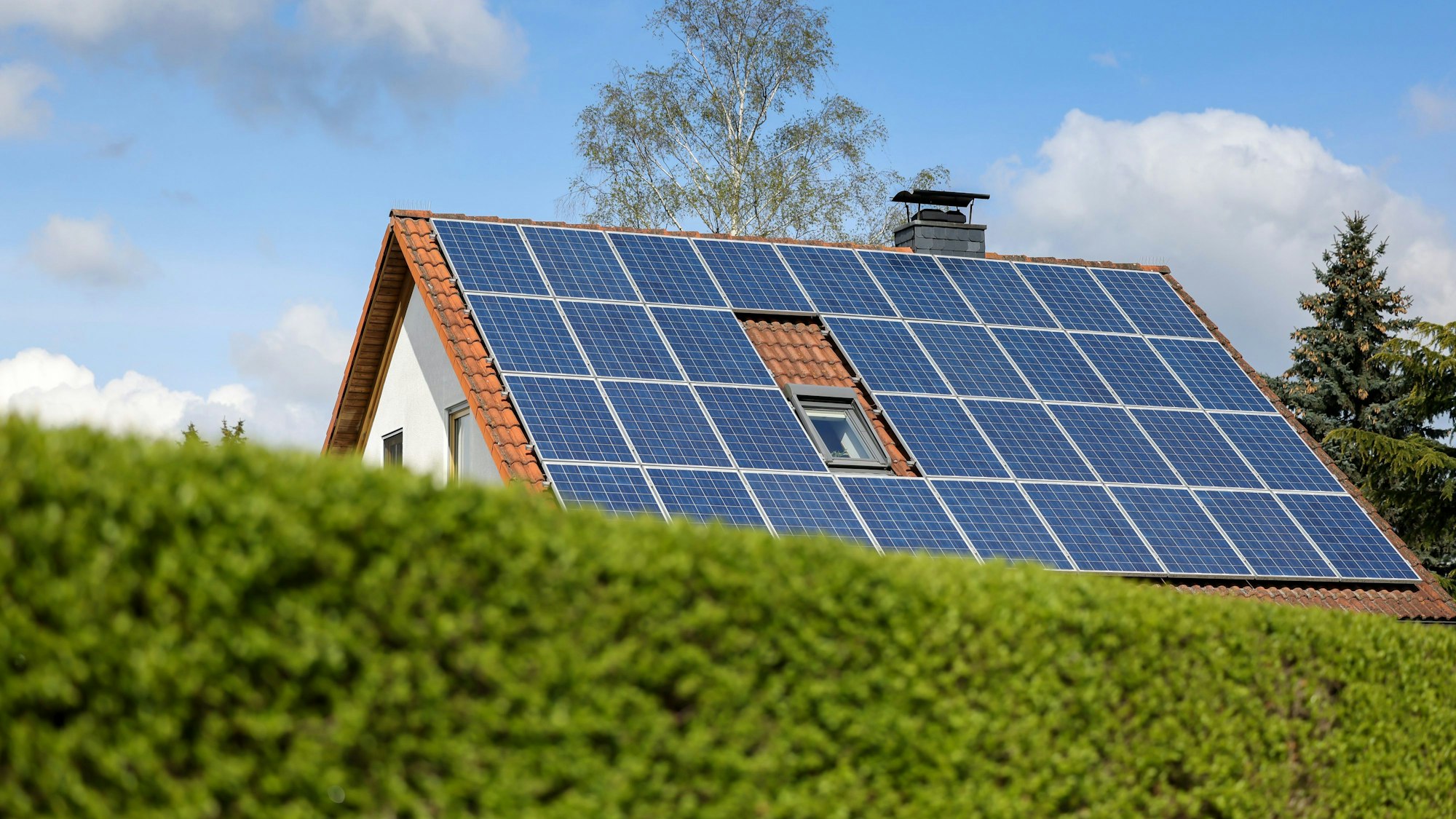 Ein Einfamilienhaus mit Solarpaneelen auf dem Hausdach steht hinter einer grünen Hecke.