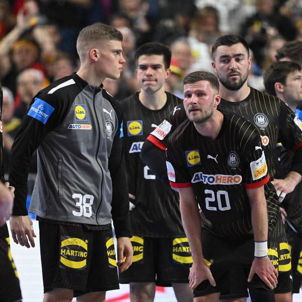 Mehrere Handballspieler stehen erschöpft und niedergeschlagen auf dem Spielfeld. Im Hintergrund befinden sich verschwommen aufgenommene Zuschauende.