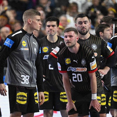 Mehrere Handballspieler stehen erschöpft und niedergeschlagen auf dem Spielfeld. Im Hintergrund befinden sich verschwommen aufgenommene Zuschauende.