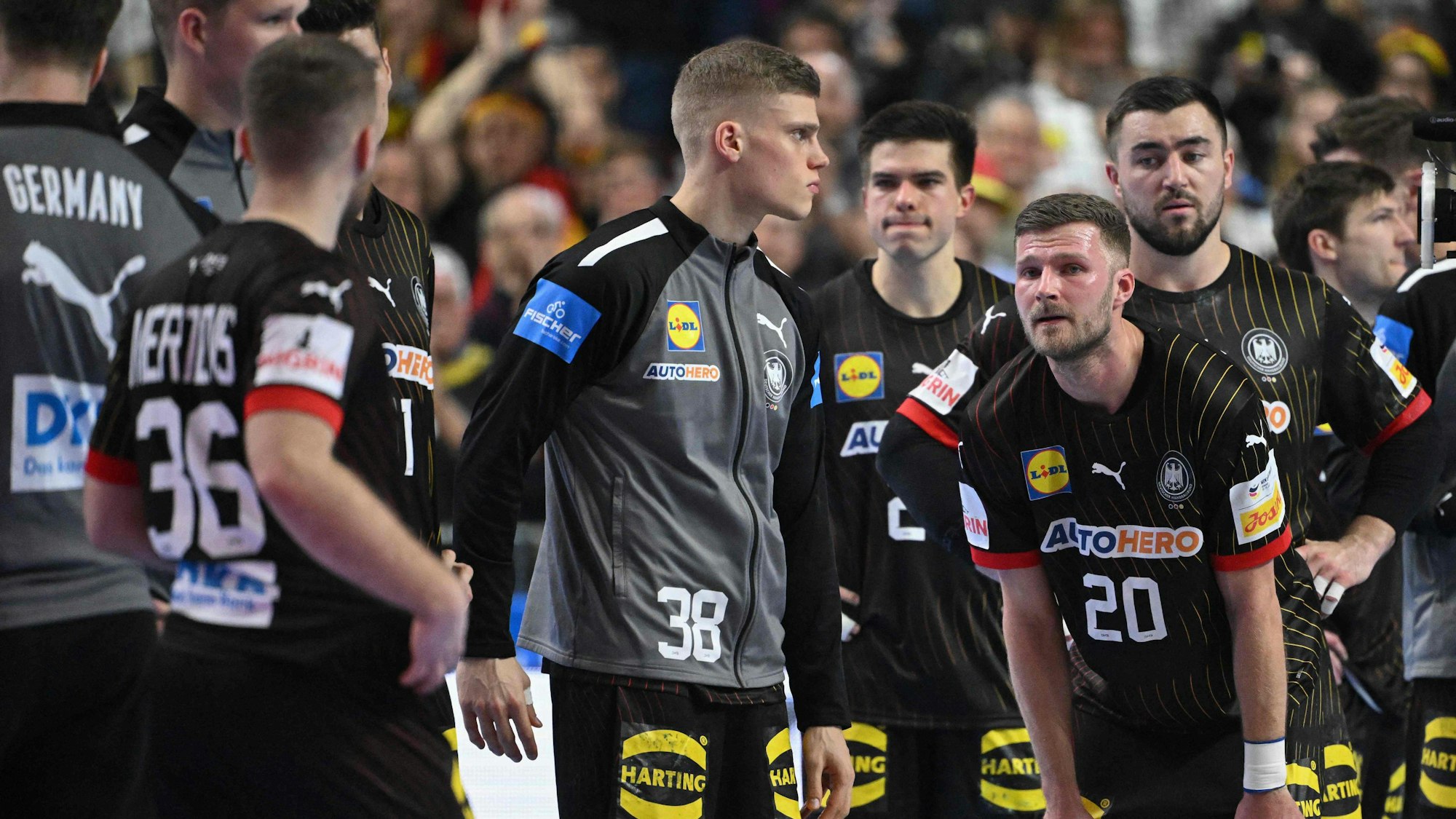 Mehrere Handballspieler stehen erschöpft und niedergeschlagen auf dem Spielfeld. Im Hintergrund befinden sich verschwommen aufgenommene Zuschauende.